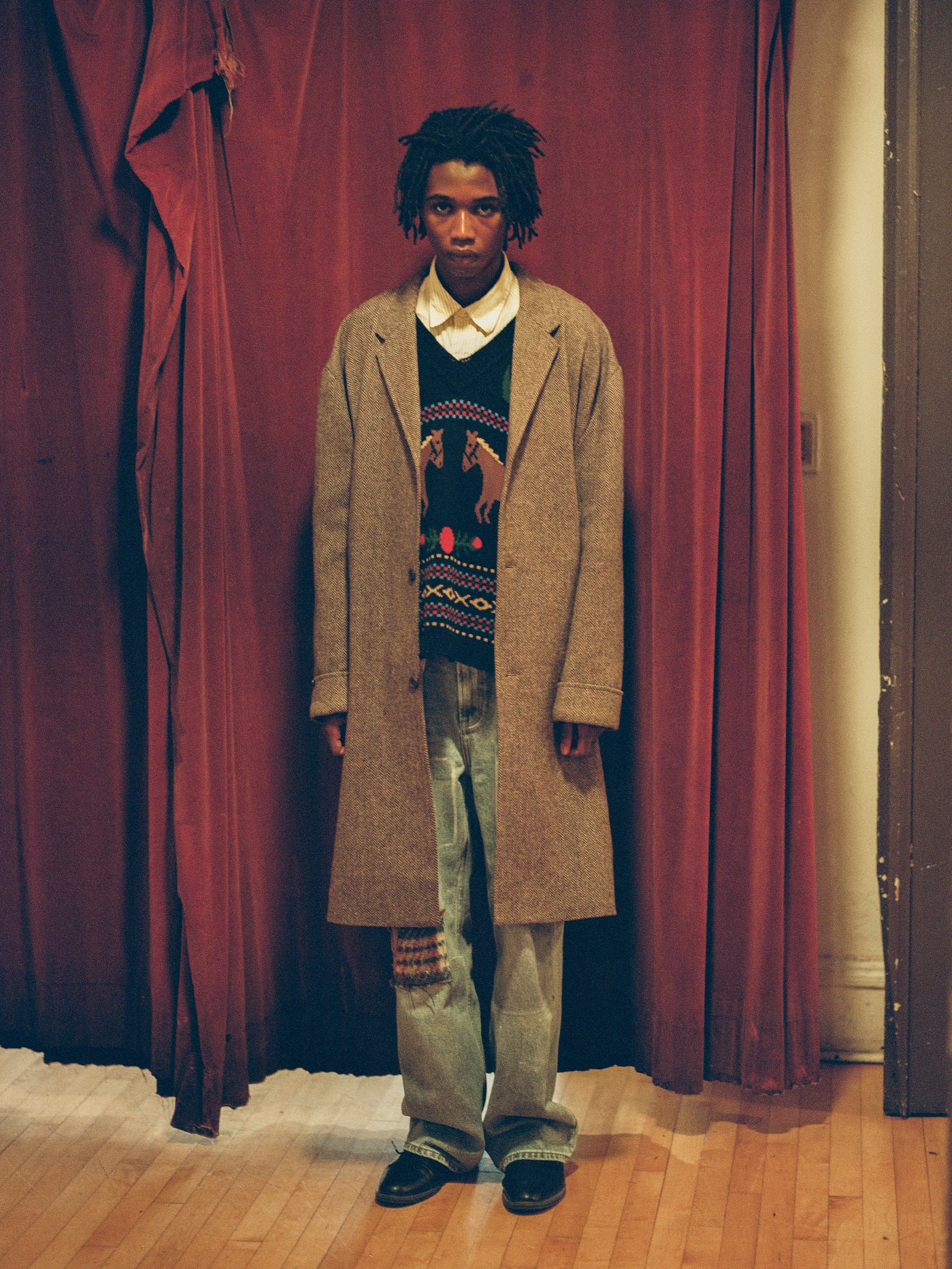 A man wearing a Found Elm Tweed Long Top Coat standing in front of a red curtain.