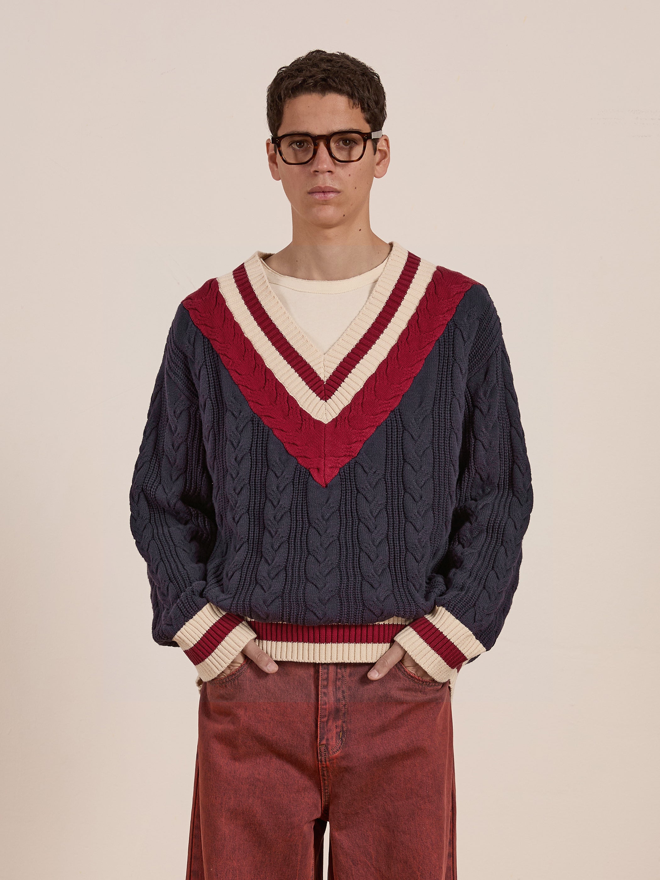 A person wearing the FOUND V-Neck Cricket Sweater in navy blue cotton with red and white cable-knit detail, along with glasses, stands against a plain background.