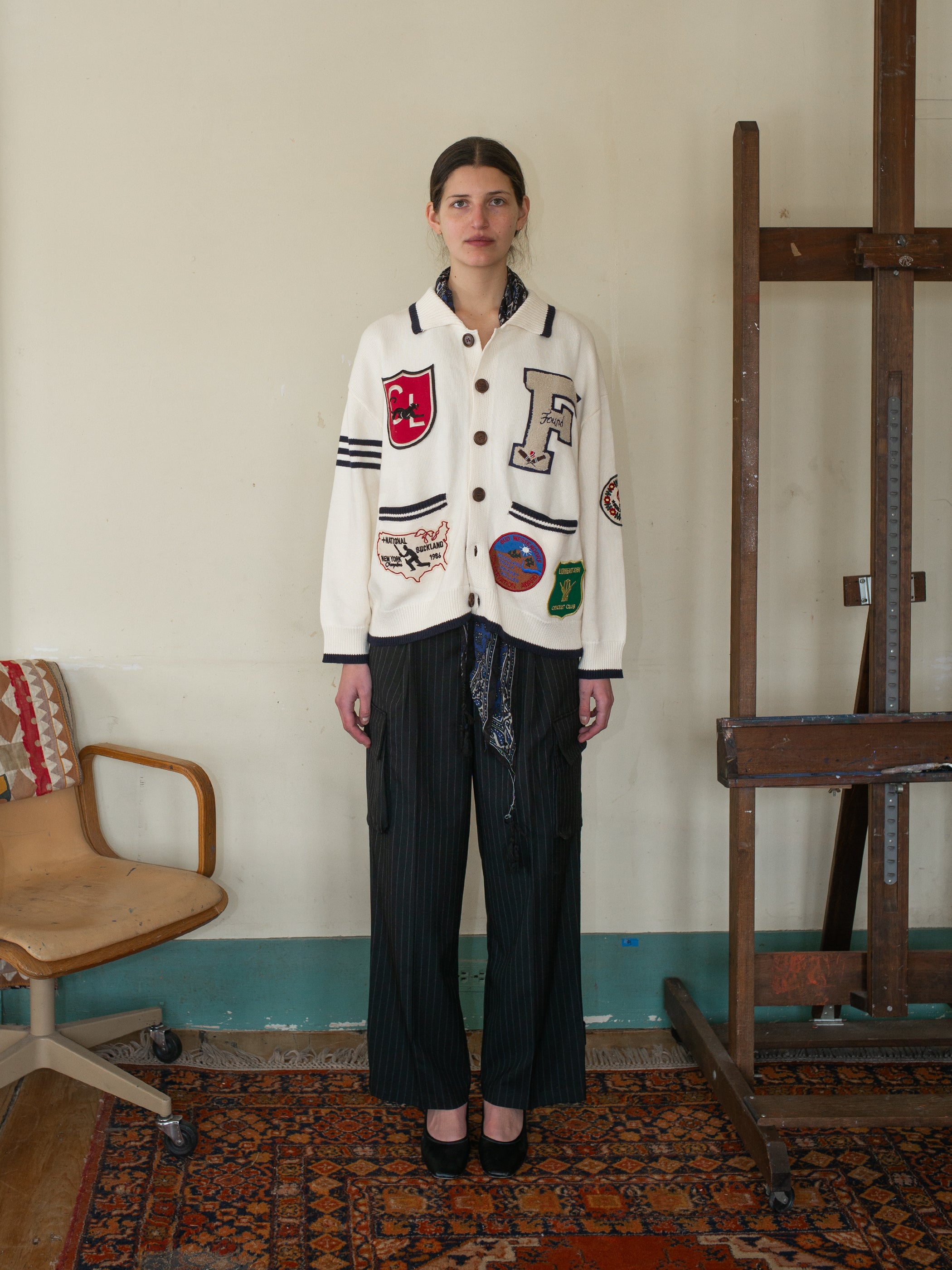 Indoors, a person dons the York Varsity Patch Collared Cardigan by FOUND along with black striped pants, creating a preppy look. An empty chair and wooden structure in the background contribute to a collegiate nostalgic vibe.