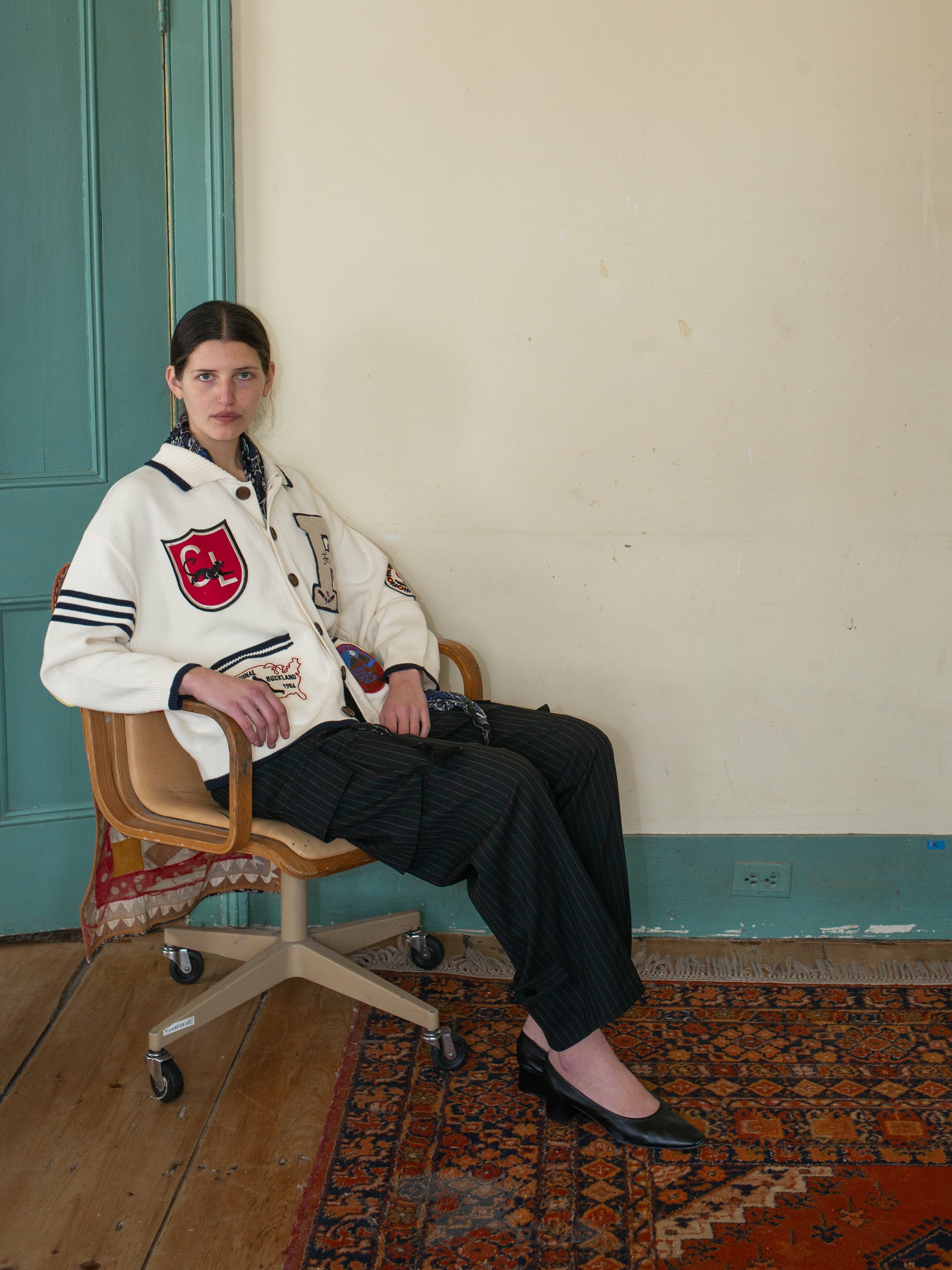 A person seated on an office chair in a room with a wooden floor and patterned rug embodies a preppy aesthetic, wearing the York Varsity Patch Collared Cardigan by FOUND, dark pinstripe pants, and black shoes against a light-colored wall.