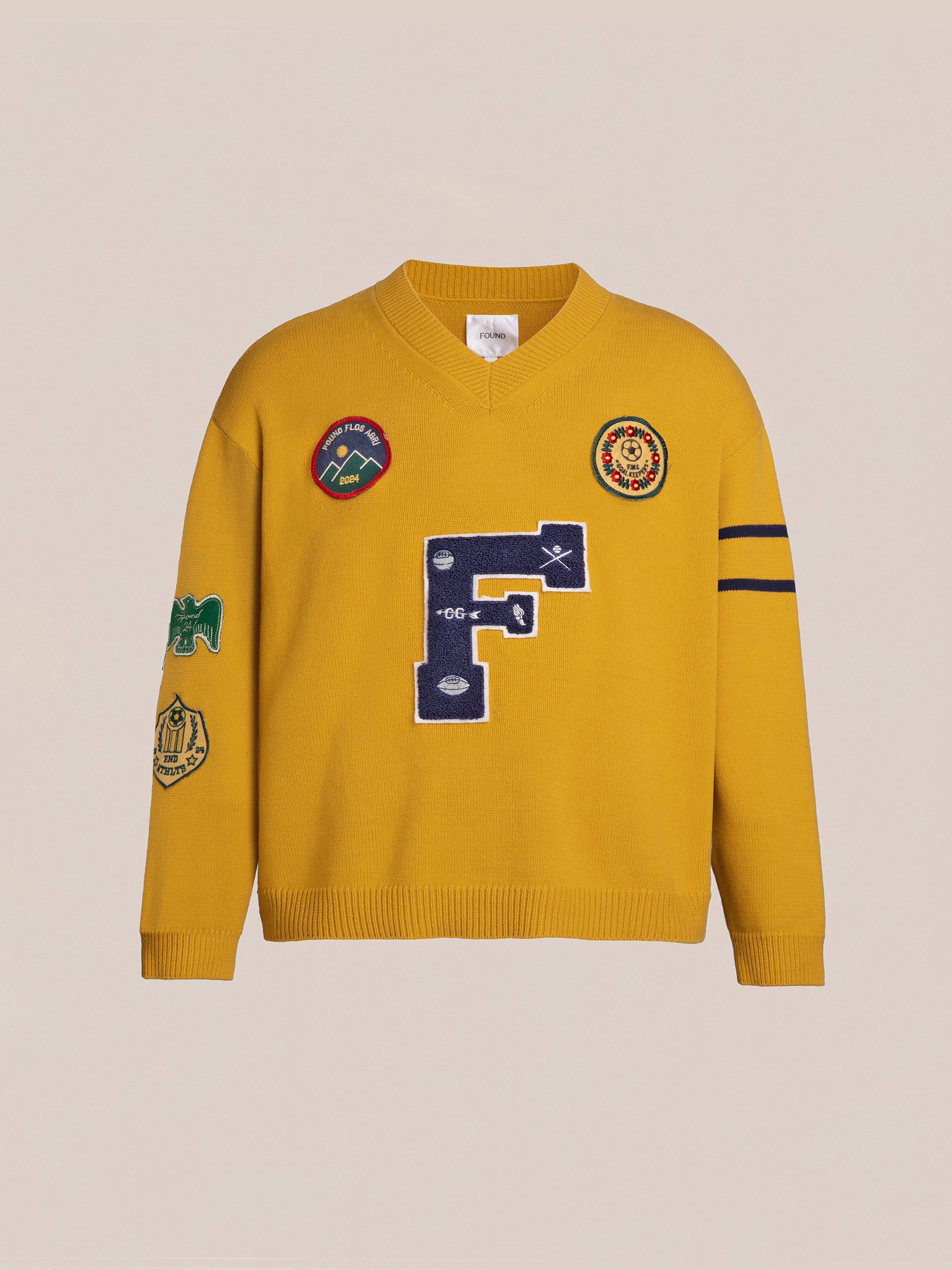 The Arrow-Wood Varsity Knit Sweater by FOUND is a yellow long-sleeve V-neck, adorned with various patches and a large blue letter 'F' on the front, perfect for adding a vintage look to your Ivy League wardrobe.