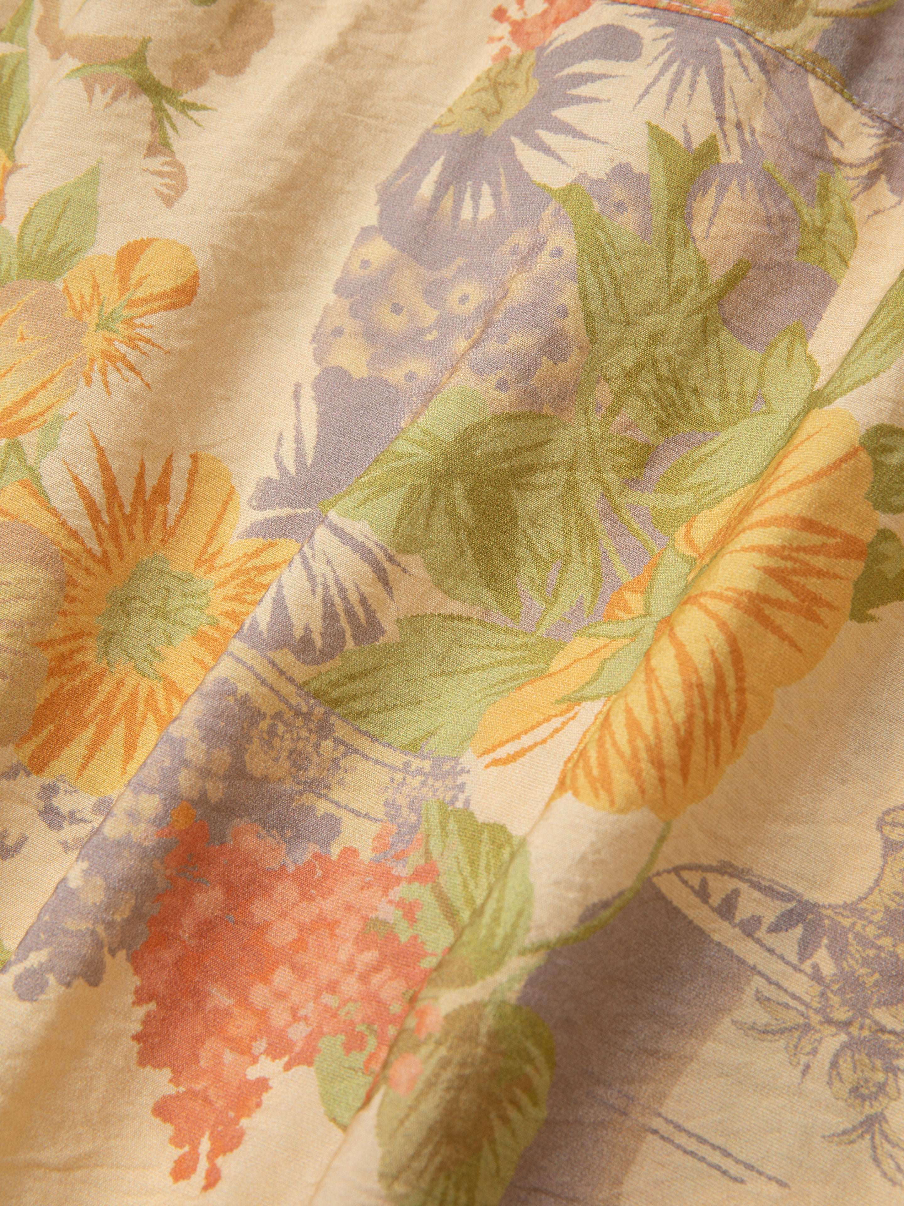 Close-up view of a fabric with a floral pattern blending sunflowers, leaves, and various other flowers in shades of yellow, orange, and green on a light background, reminiscent of traditional Phulkari motifs from Punjab Pakistan. The fabric is part of the Meraj Vase Pot Long Sleeve Camp Shirt by Found.