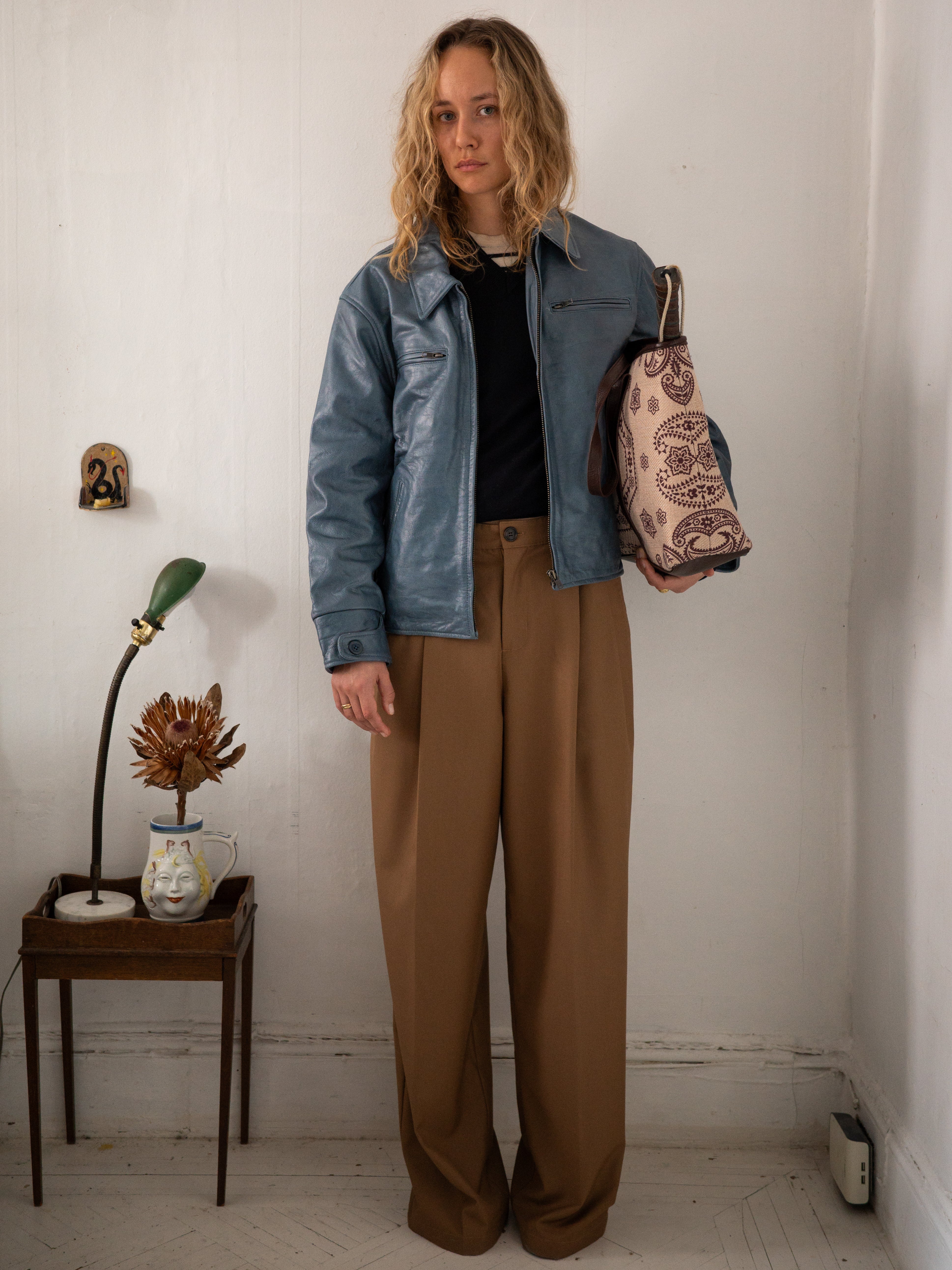 A person with long hair wears a blue leather jacket and FOUND Pleated Trousers while holding a patterned bag in a room featuring a small table and decorative items.