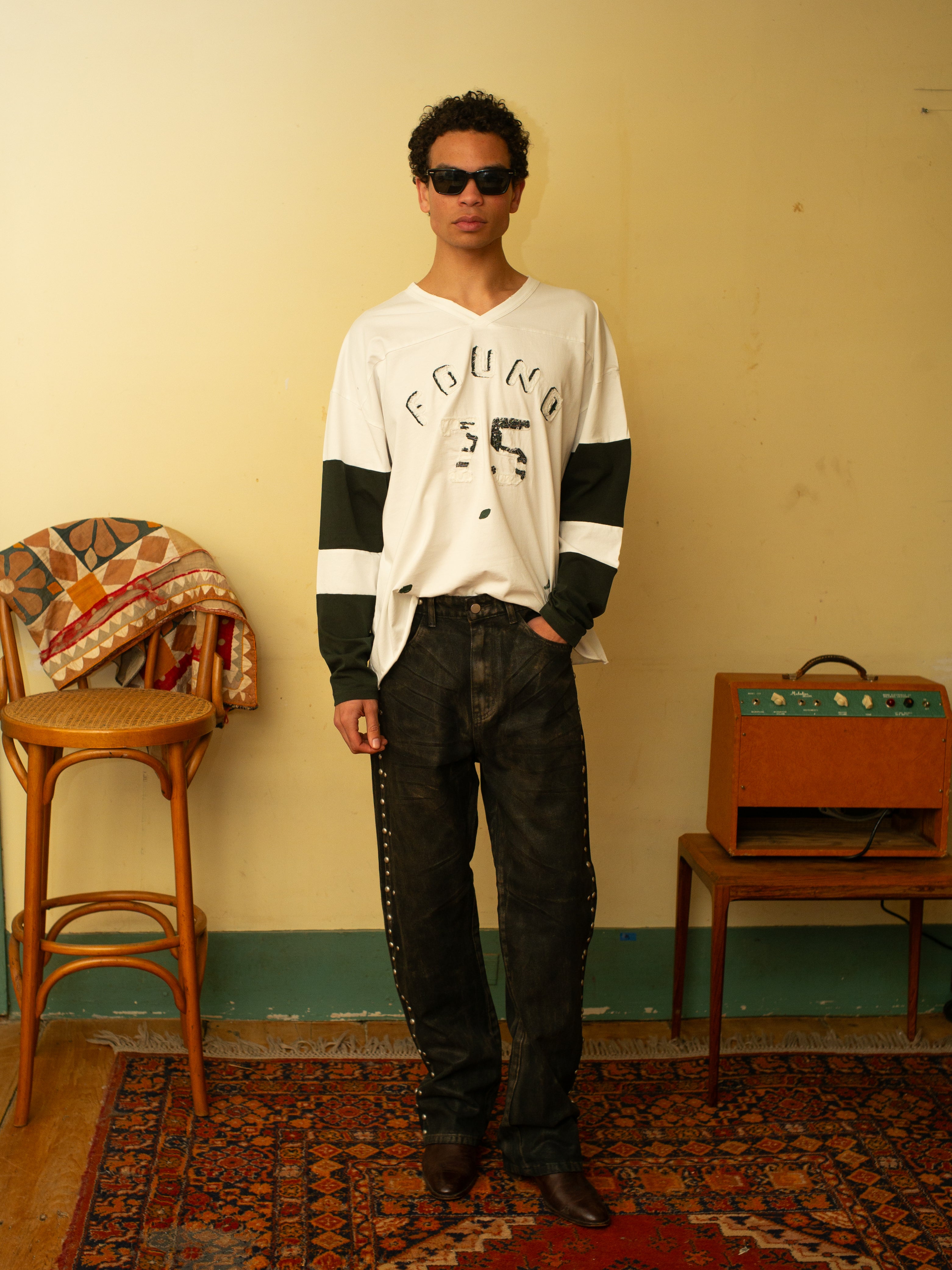 A person in sunglasses, a relaxed fit Distressed LS Team Jersey by FOUND, and jeans stands in a room with a wooden chair, quilt, and small wooden console on a patterned rug.