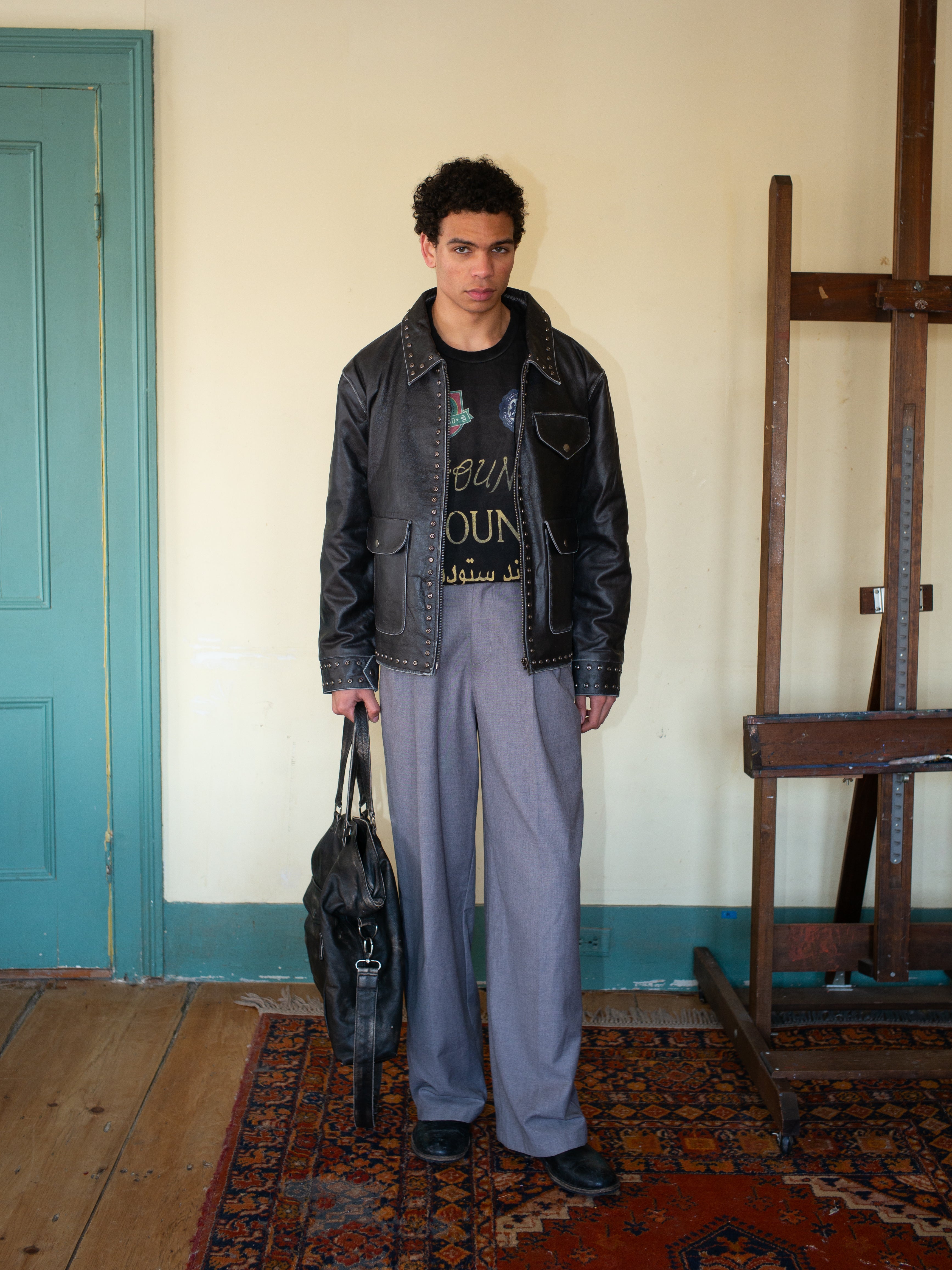 A person in a FOUND Studded Leather Pocket Jacket and gray pants stands on a patterned rug, holding a studded black bag near a wooden easel and light blue door.