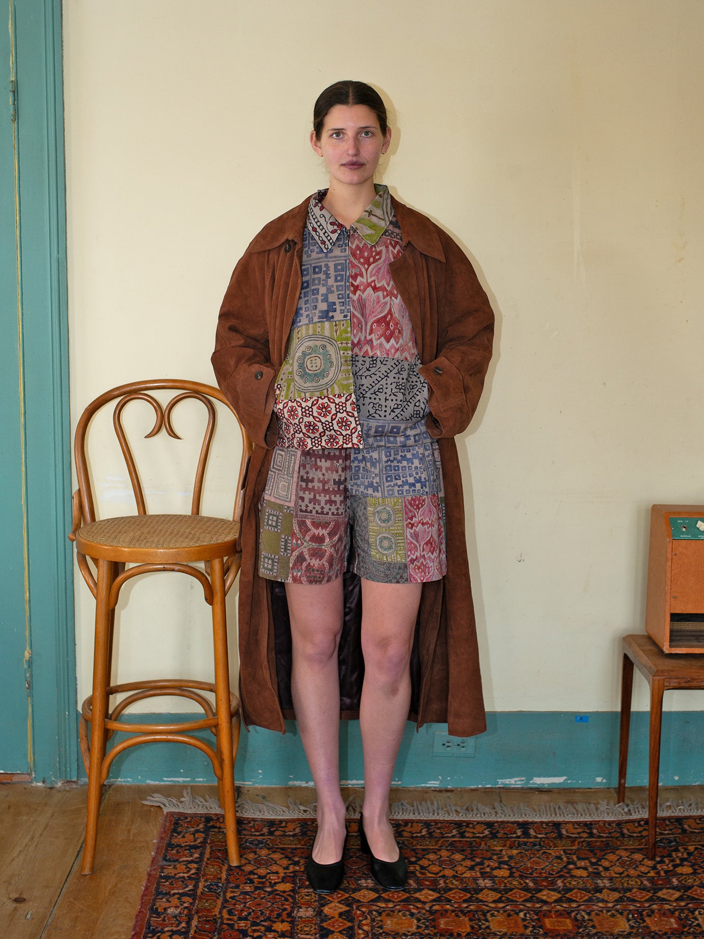 Indoors, someone wears the FOUND Watercolor Mosaic Work Jacket over a patterned dress. To their left is a wooden chair, and to the right, a small cabinet enhances the cozy ambiance.