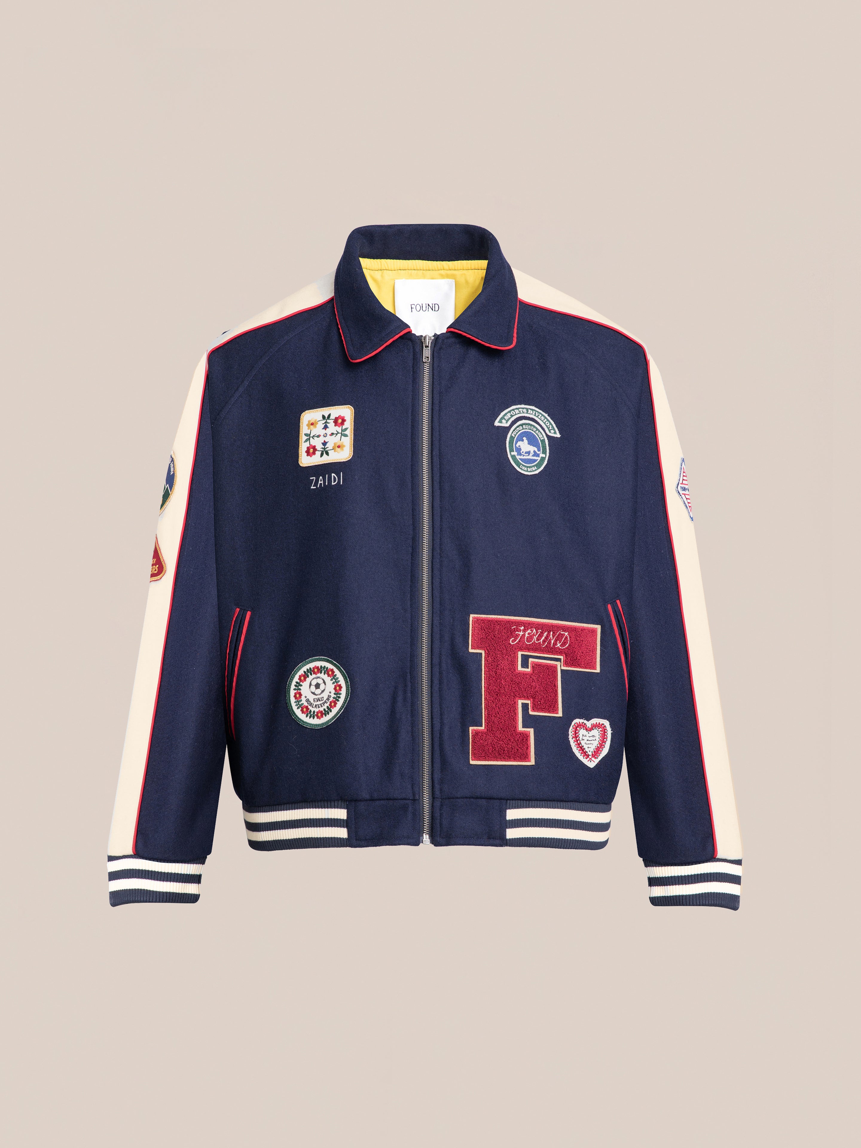 The FOUND All Roads Varsity Jacket, crafted from a mid-weight wool blend, features a navy blue design with red and white stripes on the sleeves and collar. This vintage-look jacket boasts various embroidered patches and a zip-up front.