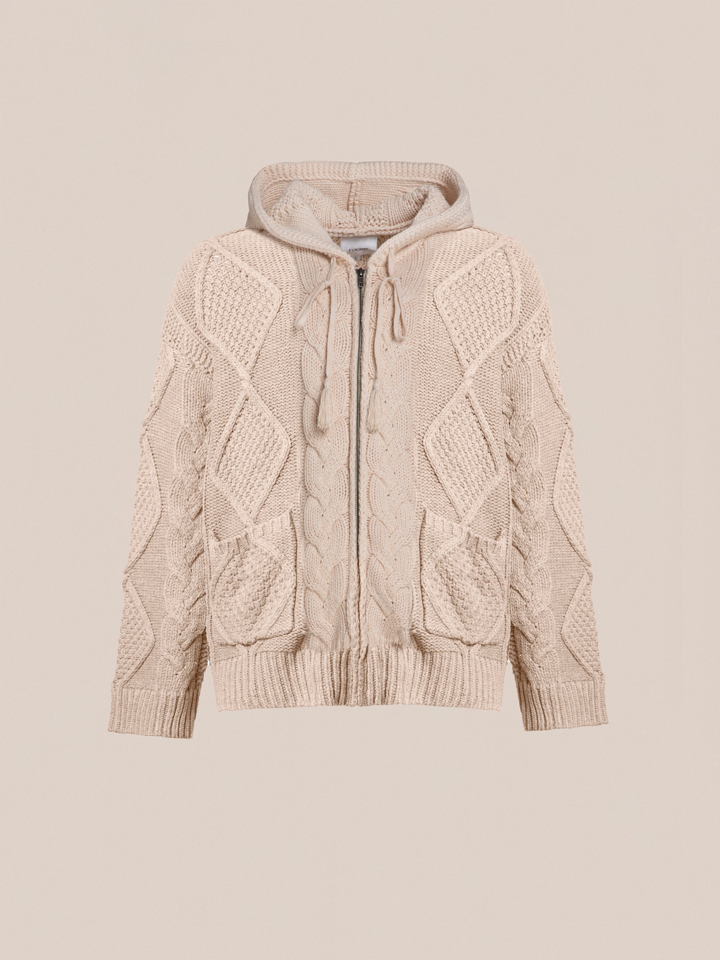 A beige, chunky knit, zip-up hoodie with a hood, featuring intricate cable knit patterns and two front pockets for comfort and warmth is the Zip Up Cable Knit Hoodie by Found.